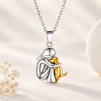 A silver pendant necklace featuring a polished silver figure hugging a small gold dog, suspended from a silver chain against a warm beige background with soft decorative props.
