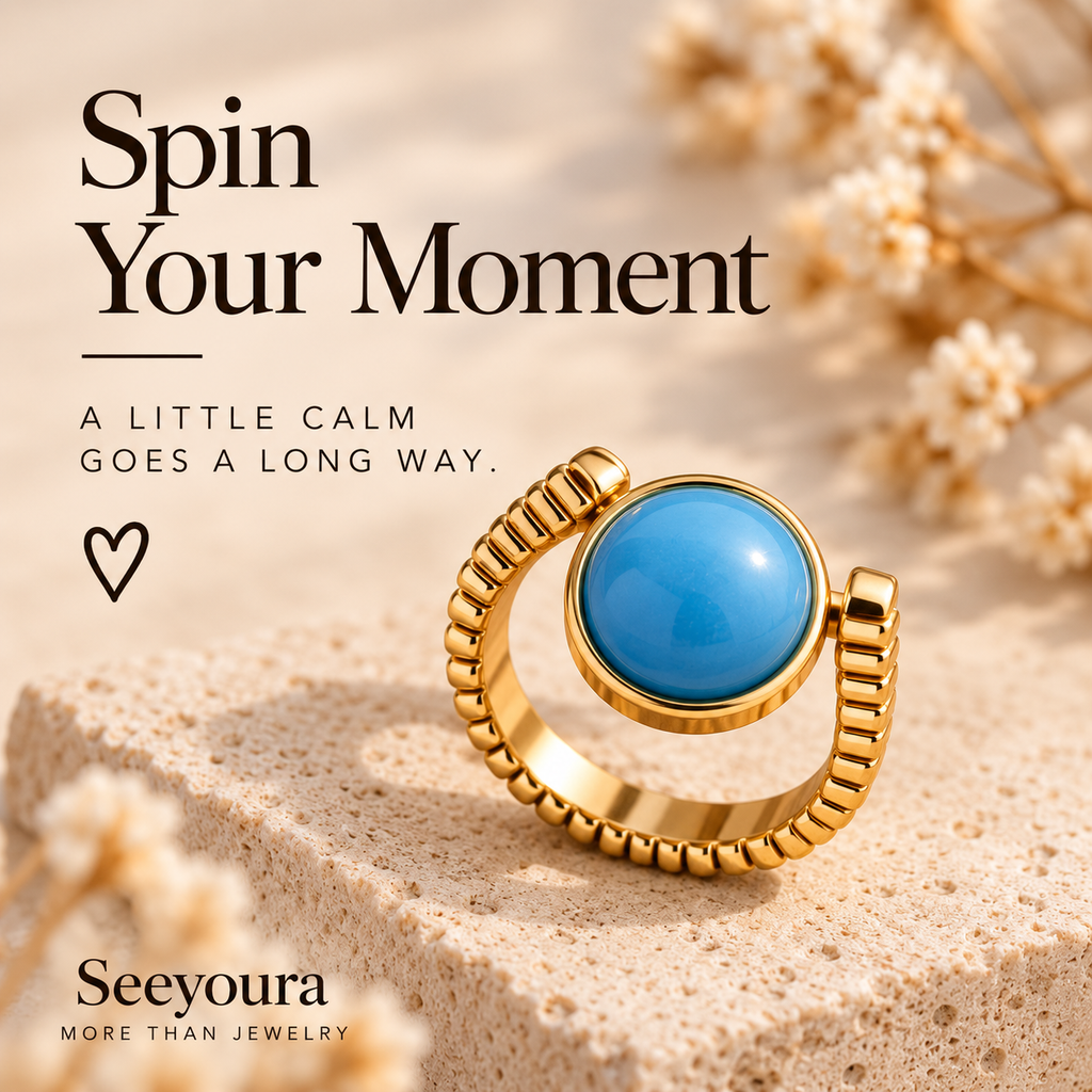 A gold ring with a round blue stone is displayed on a textured beige stone surface with dried flowers in the background. Promotional text reads 'Spin Your Moment' and 'Seeyoura MORE THAN JEWELRY'.