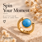 A gold ring with a round blue stone is displayed on a textured beige stone surface with dried flowers in the background. Promotional text reads 'Spin Your Moment' and 'Seeyoura MORE THAN JEWELRY'.
