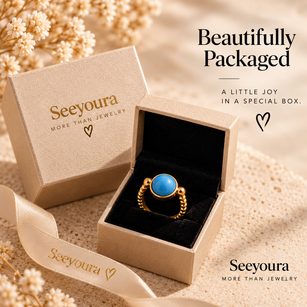 A gold ring with a round blue stone displayed in an open beige Seeyoura jewelry box, with the matching closed box and ribbon nearby on a warm beige textured surface with dried flowers. Text on the image reads 'Beautifully Packaged' and 'Seeyoura More Than Jewelry'.