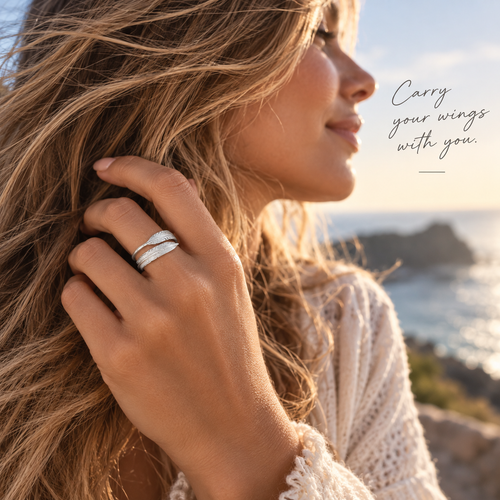 A woman in profile outdoors by the ocean at sunset, wearing two slim silver rings stacked on one finger. The image includes the text 'Carry your wings with you.