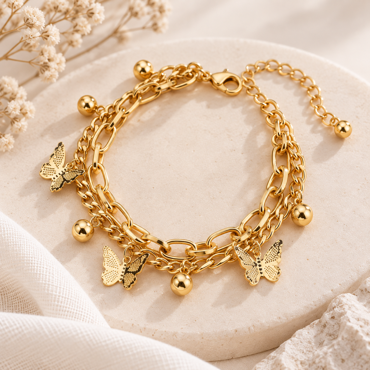 A gold-toned multi-strand chain bracelet with butterfly charms and small round bead accents, displayed on a round beige stone surface with soft fabric and dried flowers in the background.