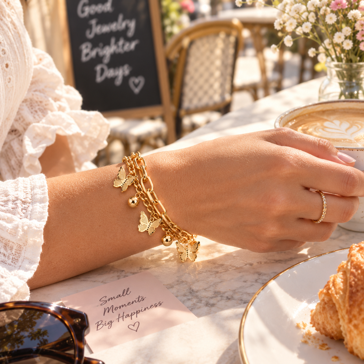 A gold charm bracelet with butterfly and bell charms is worn on a woman's wrist in a cafe setting, with a coffee cup, plate, and croissant in the background.
