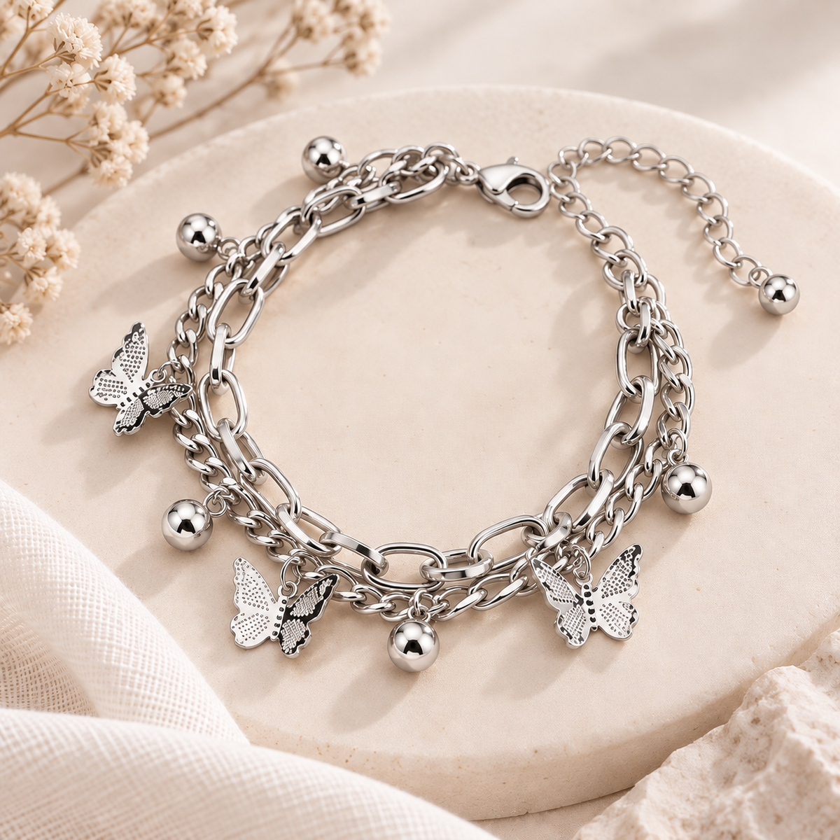 A silver-toned layered chain bracelet with butterfly charms and small bead accents, displayed on a round beige stone surface with dried flowers and soft fabric in the background.