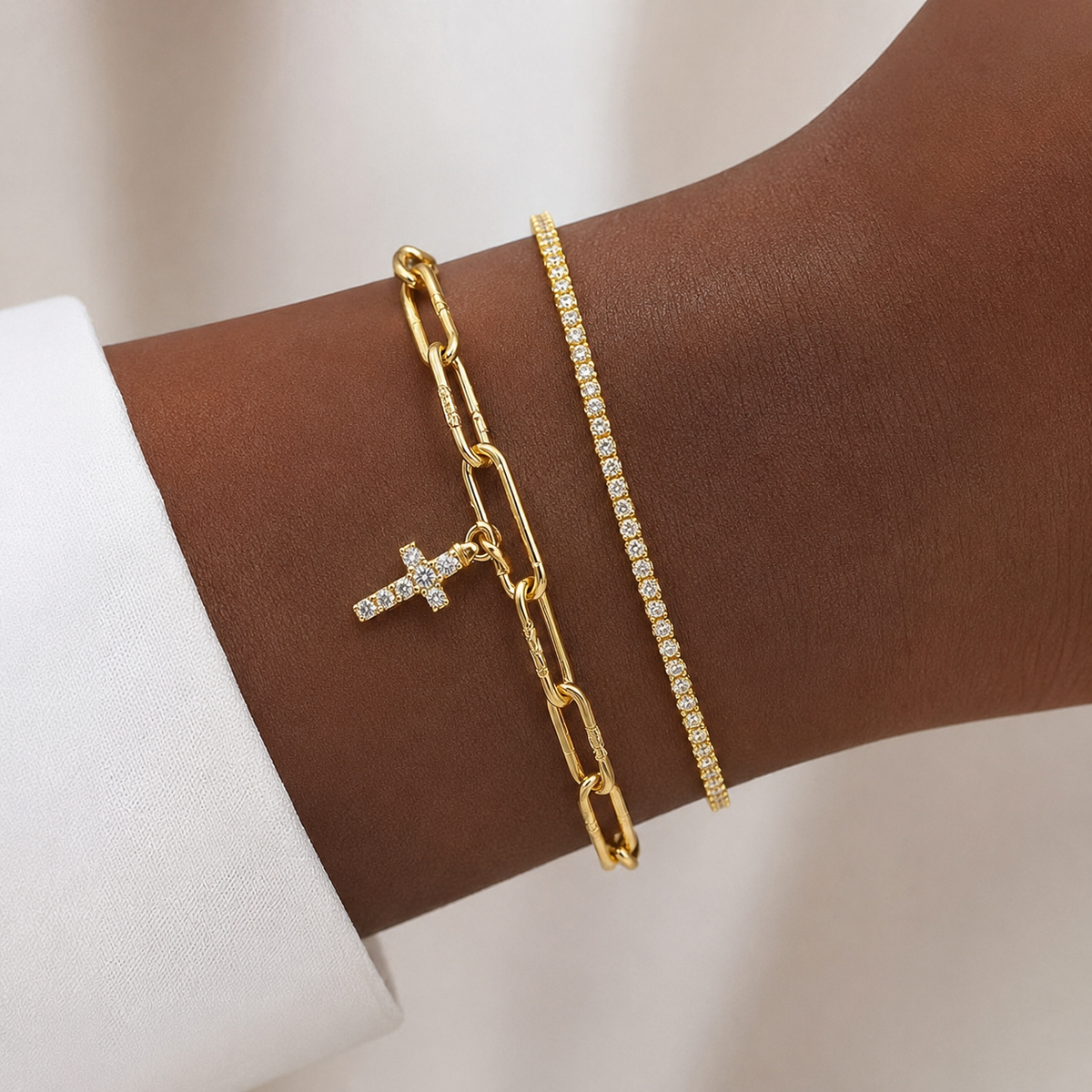 Two gold bracelets on a person's wrist: one with a cross charm set with clear stones and one with a thin line of clear stones, against a white shirt cuff and soft neutral background.