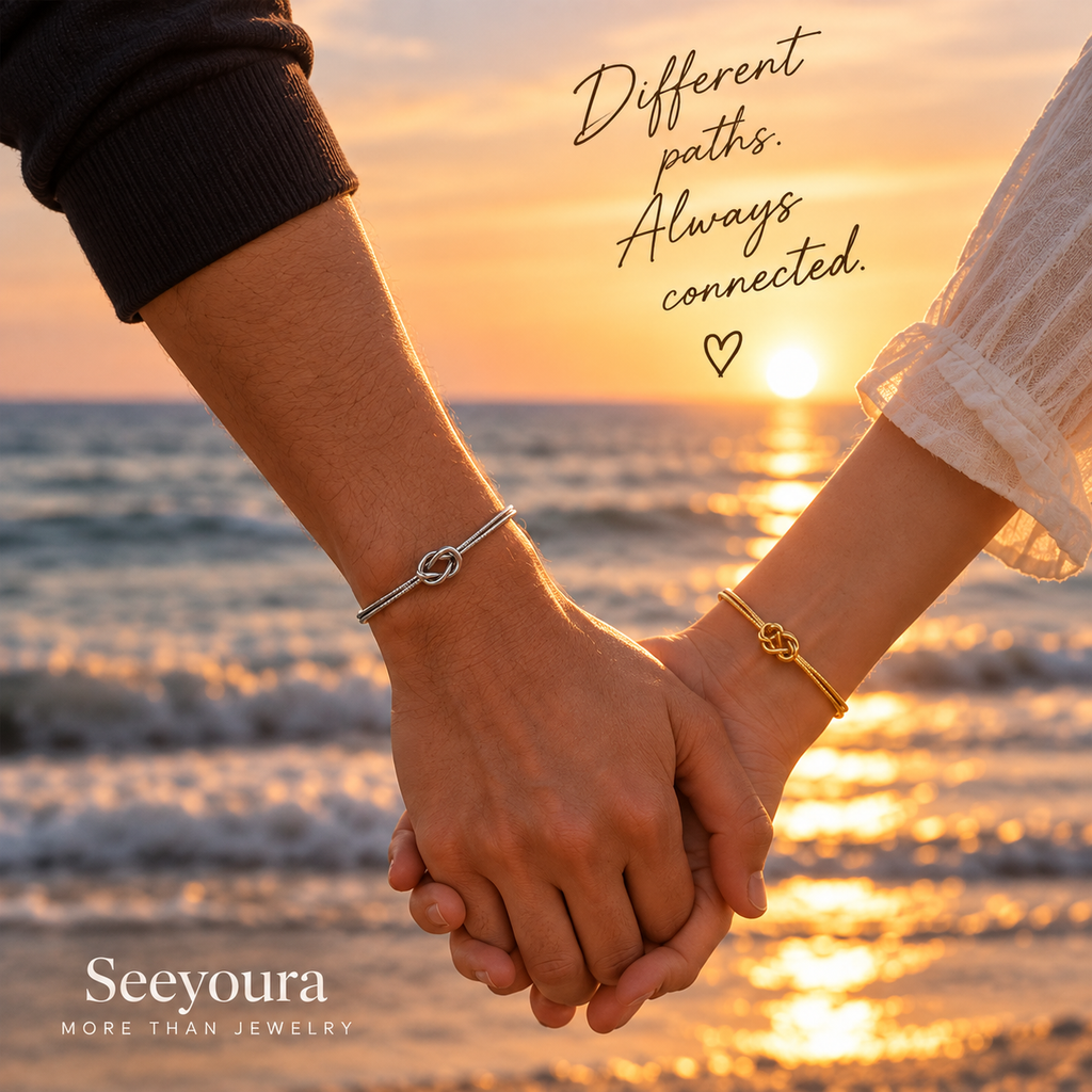 Two people holding hands on a beach at sunset, each wearing a delicate bracelet. One bracelet is silver with an infinity-style knot, and the other is gold with a matching knot.