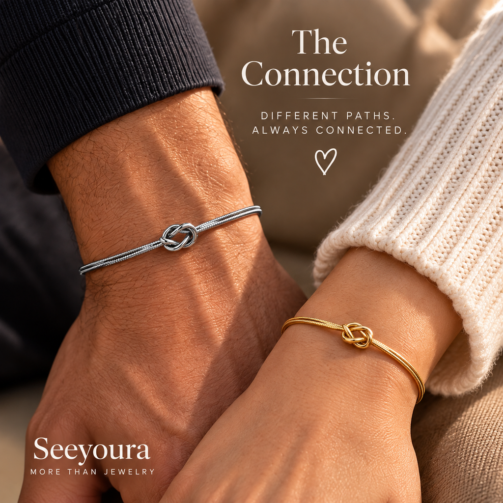 Two people holding hands, each wearing a thin bracelet with a knot design. One bracelet is silver and worn on a darker-skinned wrist, and the other is gold and worn on a lighter-skinned wrist.