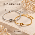 Two slim snake-style bracelets with knot centers, one silver-toned and one gold-toned, displayed on a textured beige stone surface with dried white flowers. The image includes the text 'The Connection' and 'Seeyoura MORE THAN JEWELRY', with 'S925' marks visible on the bracelet tags.